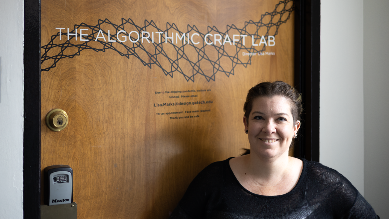 Lisa Marks standing in front of the door to her lab. The door has text saying, "The Algorithmic Craft Lab" over a stylized net pattern.