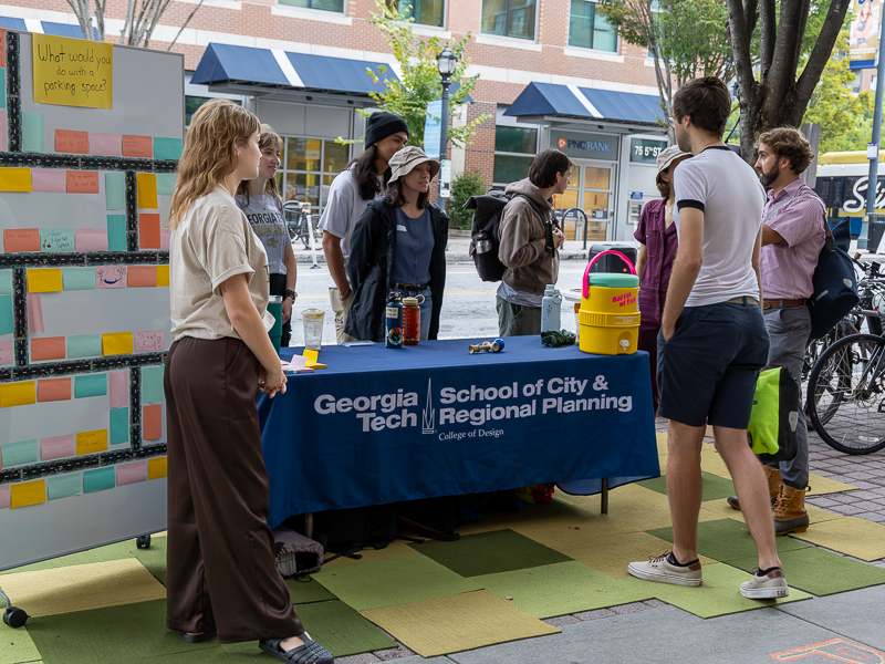 Student Planning Association members at a table outdoors talking to pedestrians. An idea board for parking space reuse stands on the left side. The tablecloth has the logo of the Georgia Tech School of City and Regional Planning.
