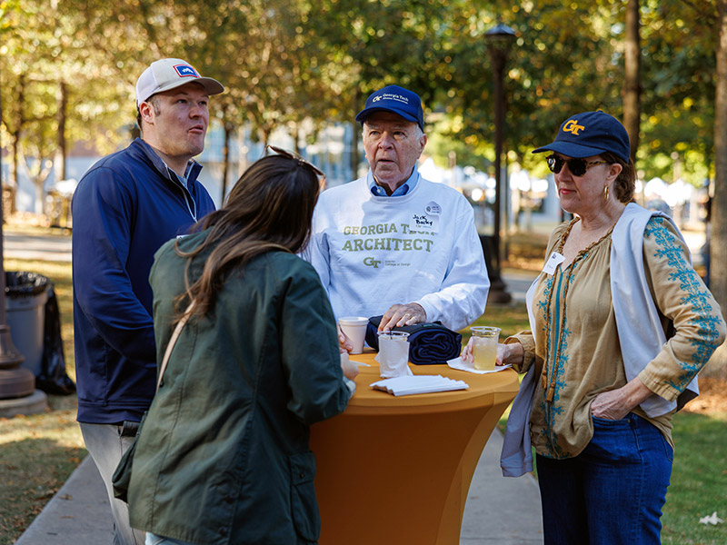 Alumni gather around a highboy table, outside on a fall day.